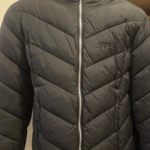 Svart pufferjacka från Svea med huva - Svart pufferjacka från Svea med snyggt broderat logotyp på bröstet och ett märke på ärmen. Säljer då jag har växt ur den!