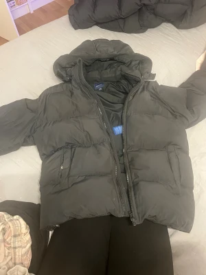 Svart pufferjacka från GANT - ordinarie pris 1999kr. Svart pufferjacka från GANT med huva och dragkedja framtill. Jackan har två fickor med lock och GANT-logga med sköld på bröstet. Insidan har en etikett för namn och telefonnummer. Perfekt för kalla dagar och riktigt snygg stil.