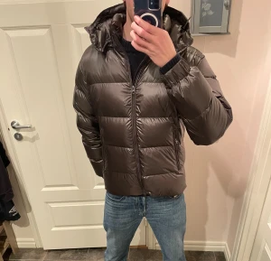 Mörkbrun/grön pufferjacka från John Henric - Säljer en mörkbrun/grön pufferjacka från John Henric med huva och dragkedja framtill. Jackan har en glansig yta. Skriv vid funderingar!!🤩