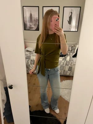 Olivgrön topp med knyt från H&M - Säljer en olivgrön topp från H&M med rund hals och korta ärmar. Toppen har en snygg knyt-detalj i sidan som ger en cool look. Materialet är mjuk bomull och passformen är normal. Perfekt att styla med jeans eller kjol. Endast använd 1 gång!!