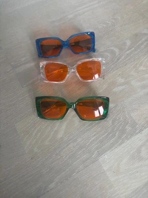 Blue-light- blocking glasögon - Tre par solglasögon med rektangulär form och transparent båge i blått, grönt och klart. Alla har orangefärgade linser som ger en cool vibe. Bågarna är tillverkade i syntetmaterial och har en modern, kantig design som sticker ut. De är för att skydda ögonen vid användning av mobilen från farligt blå ljus, de fixar sömnen och din syn. De kan också användas som solglasögon och normala glasögon för stil. De är helt nya. Köp 1 st för 50kr - köp 2 st för 85kr- köp 3 st för 110. 