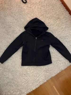Marineblå hoodie från Polo Ralph Lauren - Marineblå hoodie från Polo Ralph Lauren med röd broderad logga på bröstet. Tröjan har huva, dragkedja framtill och två fickor. Materialet är mjuk bomull och modellen är normal passform med långa ärmar. Perfekt för chill dagar och streetwear. 14-16 år 