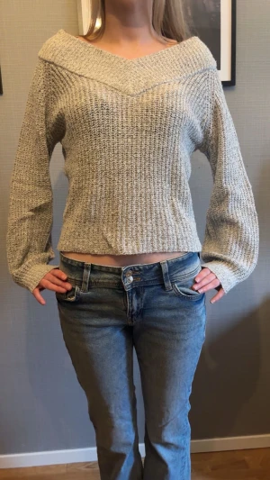 Grå stickad tröja med v-ringad krage  - Säljer en beige stickad tröja med bred v-ringad krage som går över axlarna. Tröjan har långärmade, lite lösa ärmar och ett stickat mönster. Perfekt för dig som gillar en chill och snygg stil. 