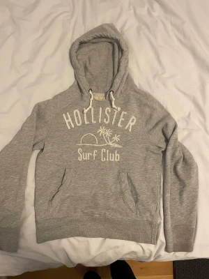 Grå Vintage Hollister Surf hoodie - Snygg grå hoodie från Hollister med broderad text 'Surf Club' och palmmotiv på bröstet. Tröjan har huva med snörning, känguruficka och ribbade muddar. Perfekt för chill dagar och har en mjuk insida i bomullsmix.
