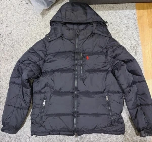 Svart pufferjacka från Polo Ralph Lauren - Svart pufferjacka från Polo Ralph Lauren med huva och dragkedja framtill. Jackan har två sidofickor med dragkedja och en röd broderad logga på bröstet. Perfekt för kyliga dagar och riktigt skön att bära.