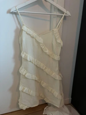 Krämvit kort klänning med volanger - Säljer en söt krämvit kort klänning med tunna axelband och v-ringad hals. Klänningen har flera diagonala volanger framtill och baktill som ger en lekfull vibe. Materialet känns lätt och luftigt, perfekt för varma dagar. Passformen är normal och klänningen är ärmlös.