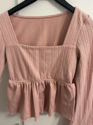 Ljusrosa ribbad blus med volang - Supersöt ljusrosa blus med ribbat mönster och bred fyrkantig urringning. Långa ärmar och volang nedtill ger en extra feminin touch. Materialet är mjukt och stickat, perfekt för dig som gillar romantisk stil och detaljer.