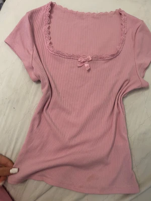 Rosa ribbad topp med spets och rosett - Supersöt ljusrosa topp med ribbad struktur, fyrkantig halsringning dekorerad med spetskant och en liten rosett framtill. Kortärmad och figurnära passform, perfekt för dig som gillar romantiska detaljer och mjuka färger.