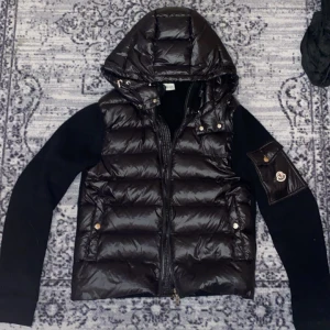 Svart dunväst från Moncler med huva - Svart dunväst från Moncler med stickade svarta ärmar och huva. Västen har quiltad framsida i glansigt material, dragkedja och fickor med tryckknappar. Moncler-logga på ärmen och snygga detaljer i guld. Perfekt för dig som gillar exklusiv streetstyle.