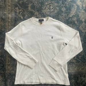 Vit långärmad tröja Polo Ralph Lauren - Vit långärmad våfflad tröja från Polo Ralph Lauren i bomull med diskret rutstruktur och klassisk broderad logga på bröstet. Tröjan har ribbade muddar och en avslappnad passform, perfekt för en clean och stilren look. Hör av er vid funderingar!