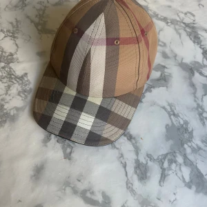 Burberry keps äkta - Säljer en klassisk keps från Burberry i deras ikoniska rutiga mönster. Kepsen är i bruna, beige och svarta toner med justerbar rem bak och broderad Burberry-logga. Tillverkad i 100% bomull med detaljer i lammnappa. Perfekt accessoar för en trendig look.