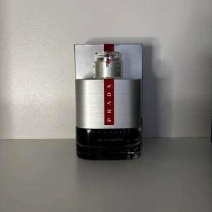 Prada Luna Rossa Eau de Toilette sampel - Snygg parfymflaska från Prada Luna Rossa med modern design i silver och svart glas. Flaskan har en röd vertikal logga och metallisk topp. Perfekt för dig som gillar exklusiva och stilrena dofter.