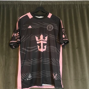 Inter Miami Messi fotbollströja L - Snygg Inter Miami fotbollströja från Adidas i svart med rosa detaljer och vågigt mönster. Messi #10 på ryggen, klubbmärke och sponsortryck på bröstet. Kortärmad och tillverkad i lätt polyester som andas. Perfekt för dig som vill sticka ut på planen eller läktaren.