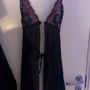 Svart spetsklänning från Chilirose - Supersöt svart kortklänning från Chilirose med transparent mesh och blommig spets upptill i svart och rosa. Djup v-ringning med glittrig detalj mellan kuporna och knytband i midjan. Perfekt för dig som gillar unika detaljer och en feminin look.