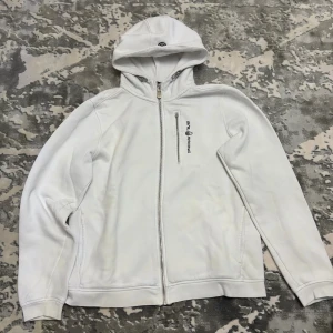 Vit hoodie från Sail Racing - Vit hoodie från Sail Racing med dragkedja framtill och broderad logga på bröstet samt på huvan. Tröjan har lång ärm, fickor i sidorna och är tillverkad i mjuk bomullsmix. Perfekt för en clean och sportig stil.