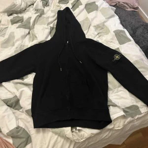 Svart hoodie från Stone Island - Svart hoodie från Stone Island med dragkedja framtill och klassisk logopatch på vänster ärm. Tröjan har huva med snörning och känguruficka.  Använd ett par gånger. Storlek S men ganska så stor i storleken