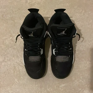 Svarta Nike Air Jordan 4 sneakers - Säljer ett par svarta Nike Air Jordan 4 sneakers med vita detaljer och klassisk Jumpman-logga på plösen och hälen. Skorna har meshpaneler, snörning och en bekväm sula med synlig Air-enhet. Perfekta för dig som gillar streetstyle och ikoniska silhuetter.