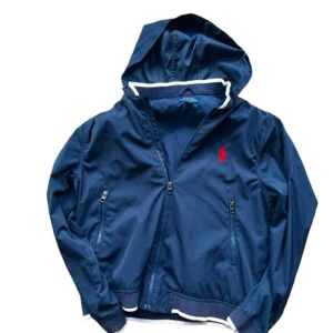 Blå windbreaker  Polo Ralph Lauren - Snygg marinblå vindjacka från Polo Ralph Lauren med vit- och marinrandiga muddar, huva och dragkedja. Jackan har två fickor med dragkedja och röd broderad logga på bröstet. Perfekt för dig som gillar klassisk och sportig stil. Nypris ca 2500kr; vårt pris 599! Priset är inte hugget i sten🙌Storleken är L i junior vilket motsvarar S. 
