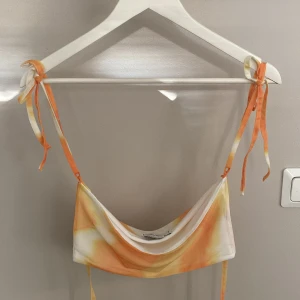 Orange och vit topp - NA-KD - Superfin topp från Angelica Blick x NA-KD i storlek S. Snyggt mönster i orange, gul och vit med tunna axelband som knyts på axlarna. Bandeau-modell med draperad framsida, perfekt för sommarens strandhäng.