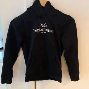 Svart hoodie Peak Performance 140 - Svart hoodie från Peak Performance med broderad vit logga på bröstet. Klassisk känguruficka framtill, huva och långa ärmar. Perfekt för chill dagar eller när du vill ha en enkel och snygg look.