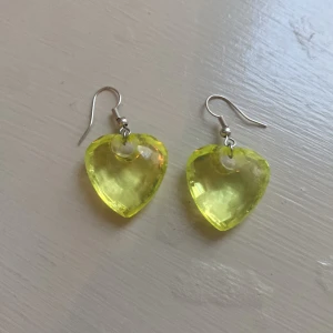 Gula hjärtformade örhängen - Säljer ett par coola örhängen med stora, transparenta hjärtan i klar gul färg. Perfekta för att ge din outfit en färgklick och lite extra attityd. Krokar i silverfärgade metalldelar. Superfina till både vardag och när du vill sticka ut.