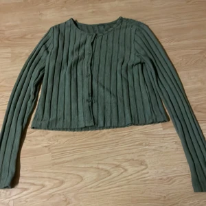 Olivgrön ribbad kofta med knappar - Snygg grön kofta med ribbad struktur och långa ärmar. Koftan har rund halsringning och knäppning framtill med matchande knappar. Perfekt att slänga över en topp för en chill look.