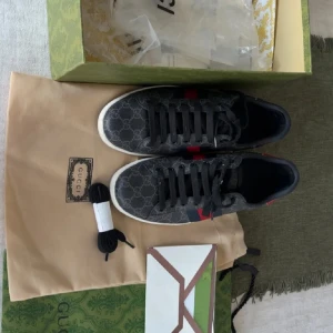 Gucci sneakers med GG-mönster - Säljer ett par svarta Gucci sneakers med klassiskt GG-monogram i canvas, röda och blå ränder på sidorna och metallicröd samt metallicblå häl med ormskinnslook. Skorna har svarta snören och vit sula. Kommer med originalkartong, dustbag och extra skosnören.