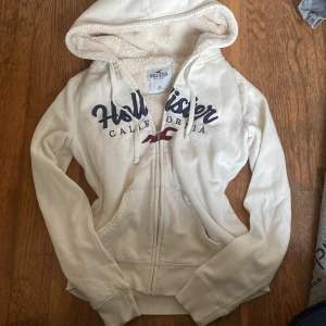Créme vit hoodie från Hollister  - Mysig vit hoodie från Hollister med blå broderad logga och dragkedja framtill. Fodrad med fluffigt teddyfoder. Passar perfekt till hösten och vintern nu!