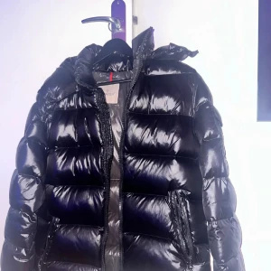 Moncler maya pufferjacka - Svart glansig pufferjacka från Moncler med huva och klassisk logga på ärmen. Jackan har dragkedja framtill, två sidofickor med dragkedja och ett coolt serietryck på insidan. Perfekt för dig som vill ha en varm och snygg vinterjacka med streetkänsla.
