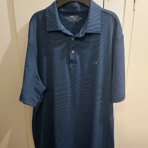 Blå randig polo från Vineyard Vines - Snygg blå kortärmad polo från Vineyard Vines med tunna vita ränder och klassisk krage. Tröjan har två knappar framtill och en liten broderad val på bröstet. Tillverkad i mjuk polyester och spandex för skön stretch och passform.