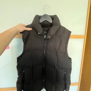 Armani Väst - Svart dunväst från Armani Jeans med hög krage och två dragkedjeförsedda fickor. Västen har quiltad design och stängs med dragkedja framtill. Perfekt för lager-på-lager och har en clean, stilren look som funkar till många outfits.