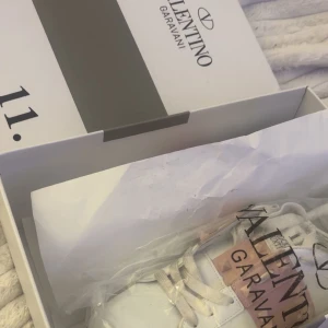 Valentino Rockstud sneakers vit/rosa - Säljer ett par Valentino Garavani Rockstud sneakers i vitt skinn med ljusrosa band och ikoniska nitar. Skorna har snörning och platt sula, samt rund tå. Märkeslogga på plösen och insidan. Perfekta för dig som vill ha en lyxig och edgy look.