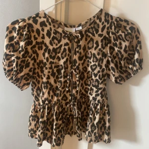 Gina tricot Leopardmönstrad blus med puffärm - Säljer en snygg blus från Gina tricot i leopardmönster. Toppen har korta puffärmar, volang nertill och knytning framtill. Färgerna är beige och svart, och materialet känns mjukt och lätt. Perfekt för dig som vill sticka ut med ett djurmönster!