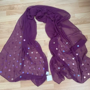 Ny.Lila sjal med paljetter - Snygg lila sjal i tunn, transparent polyester med glittriga paljetter i olika nyanser. Halsduken är lång och luftig, perfekt att svepa runt halsen för en cool och färgstark look. Paljetterna ger extra shimmer och gör den till ett statement-plagg. 
