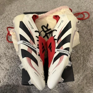 Adidas Predator Elite FT FG fotbollsskor - Säljer ett par Adidas Predator Elite FT FG fotbollsskor i vitt, svart och rött. Skorna har snörning, tydliga svarta ränder och röda detaljer, samt texten 'PREDATOR' under sulan. Designade för gräsplan med fasta dobbar och tillverkade i syntetmaterial.