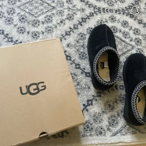 Svarta UGG boots med fårfoder - Mysiga svarta UGG boots i mocka med fluffigt fårfoder på insidan. Snyggt broderad kant i vitt och grått runt öppningen. Klassisk rund tå och platt sula med UGG-logga undertill. Perfekta för kalla dagar när du vill hålla fötterna varma.