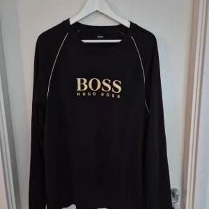 Svart tröja från Hugo Boss - Svart långärmad tröja från Hugo Boss med guldfärgad logga framtill och tunna guldiga sömmar längs axlarna. Klassisk rund halsringning och mjukt material som känns skönt mot huden. Perfekt för en stilren och avslappnad look.