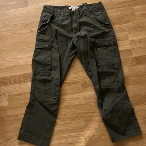 Mörkgröna cargobyxor med fickor - Snygga mörkgröna cargobyxor med flera stora fickor på sidorna och bak. Byxorna har rak passform och är tillverkade i ett slitstarkt bomullstyg. Perfekta för dig som gillar en avslappnad och funktionell stil.