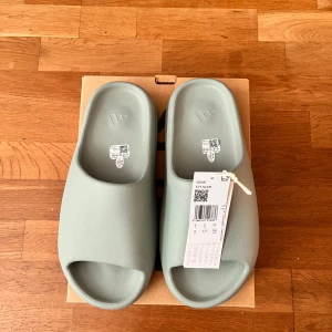Adidas Yeezy Slide salt - Adidas Yeezy Slide i en fräsch ljusgrön färg. Slidesen har en chunky, ribbad sula och öppen tå. Tillverkade i syntetmaterial för en clean och modern look. Perfekta för chill dagar och enkel att slänga på sig.