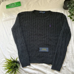 Mörkblå kabelstickad tröja Polo Ralph Lauren - Mörkblå kabelstickad tröja från Polo Ralph Lauren med lila broderad logga på bröstet. Tröjan har rund halsringning, ribbade muddar och är långärmad. Perfekt för dig som gillar klassisk och stilren look.