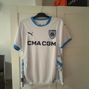 Marseille vit matchtröja Puma - Snygg vit fotbollströja från Puma för Olympique de Marseille med blå detaljer vid krage och ärmslut. Tryckt klubbmärke och sponsorlogga på bröstet. Unika ljusblå mönster längs sidorna. Perfekt för dig som vill representera din klubb.