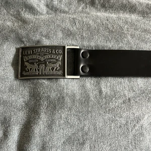 Svart skärp från Levi's i skinn - Snyggt svart bälte från Levi's i äkta skinn med en robust metallspänne som har klassisk Levi Strauss & Co-gravyr. Perfekt accessoar för att lyfta din outfit med en tidlös och stilren look.