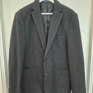 Mörkgrå kavaj Dressmann slim fit - Snygg mörkgrå kavaj från Dressmann i slim fit-modell. Kavajen har två knappar framtill, tre utanpåfickor och klassisk krage. Fodret har ett diskret blommönster. Perfekt för dig som vill ha en stilren och modern look.