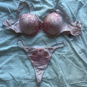Rosa blommigt set med broderi - Supersöt bh-topp och matchande trosor i ljusrosa med broderade blommor i rosa och guld. Kuporna är vadderade och dekorerade med mesh och små rosetter framtill. Materialet känns mjukt och lyxigt, perfekt för dig som gillar romantiska detaljer och blommigt mönster. OANVÄNT!!!! Storlek L/XL