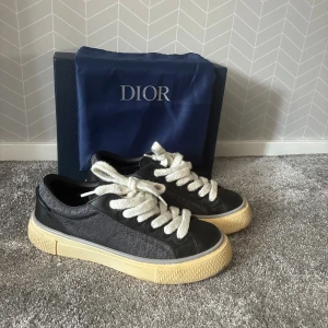 Dior sneakers - Säljer ett par svarta Dior sneakers med diskret monogrammönster på sidorna, tjocka vita snören och beige sula. Skorna har en klassisk låg profil och är tillverkade i en mix av skinn och textil. Perfekta för dig som vill ha en lyxig och stilren look. Dustbag och boxen tillkommer vid köp. Kvitto finns som autentiskt bevis!