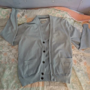 Ljusblå kofta från GANT - Snygg ljusblå kofta från GANT med klassisk v-ringning och svarta knappar framtill. Koftan har två fickor fram och är tillverkad i mjuk bomull. Perfekt för lager-på-lager och chill stil. Långärmad och passar till många outfits. 98% bommull och 8% kashmir