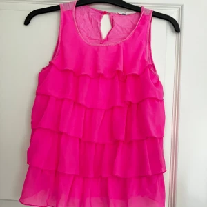 Rosa volangtopp från H&M - Superfin ärmlös topp från H&M i stark rosa färg med flera lager volanger framtill. Toppen har rund halsringning och en liten knappöppning bak i nacken. Perfekt för dig som vill sticka ut med en färgglad och lekfull stil.