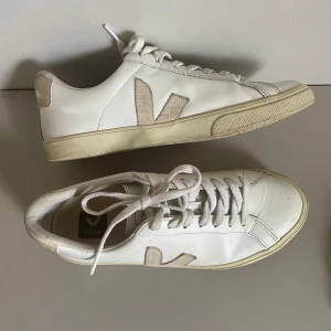 Vita Veja sneakers med beige detaljer - Snygga vita sneakers från Veja med beige mockadetaljer och klassisk logga på sidan. Skorna har rund tå, snörning och platt sula. Ovandelen är i skinn och detaljer i textil och mocka. Perfekta för en clean och stilren look. Använda mycket så har en liten defekt på framsidan av höger fot. Nypris 1700kr. 