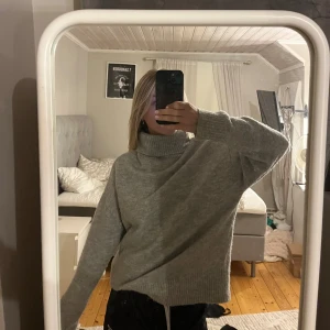 Grå polotröja i stickat material - Mysig grå polotröja i stickat material med oversized passform och långa ärmar. Tröjan har en hög, ribbad polokrage och breda muddar vid ärmslut och nederkant. Perfekt för lager-på-lager under kyliga dagar.