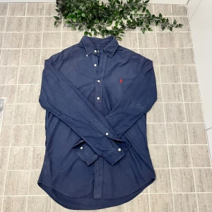 Mörkblå skjorta Ralph Lauren slim fit - Klassisk mörkblå skjorta från Ralph Lauren i slim fit-modell. Skjortan har långa ärmar, knappar framtill och ikonisk röd broderad logga på bröstet. Tillverkad i mjuk bomull och passar perfekt till en stilren look.
